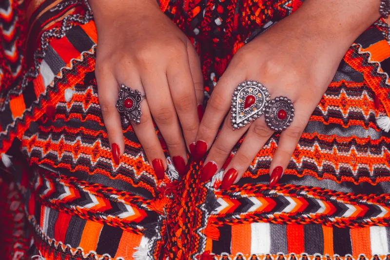 Hands with red nail polish and silver rings resting on embroidered fabric